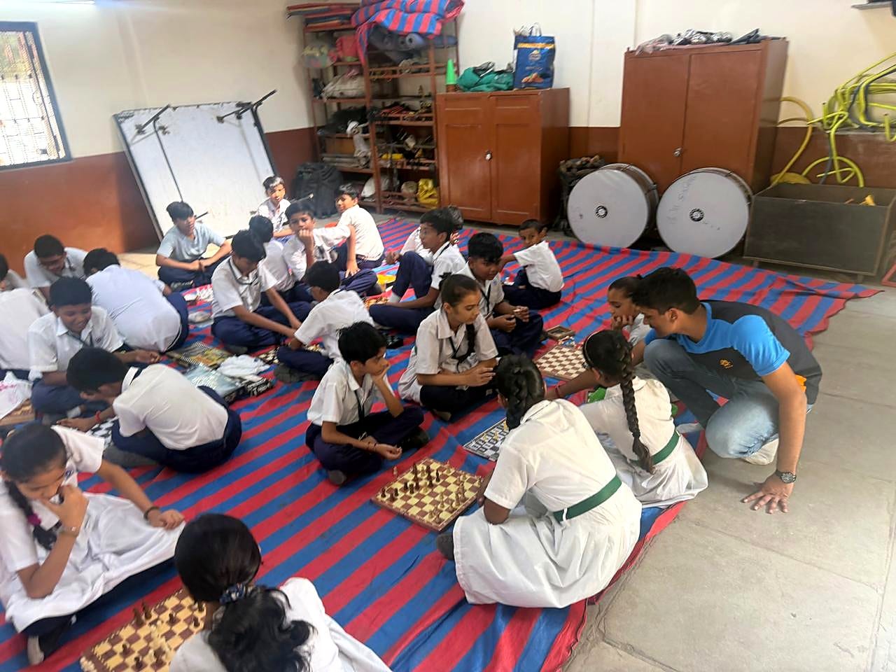 Students practise in small groups on a striped mat while Mukund guides a group at a chessboard.