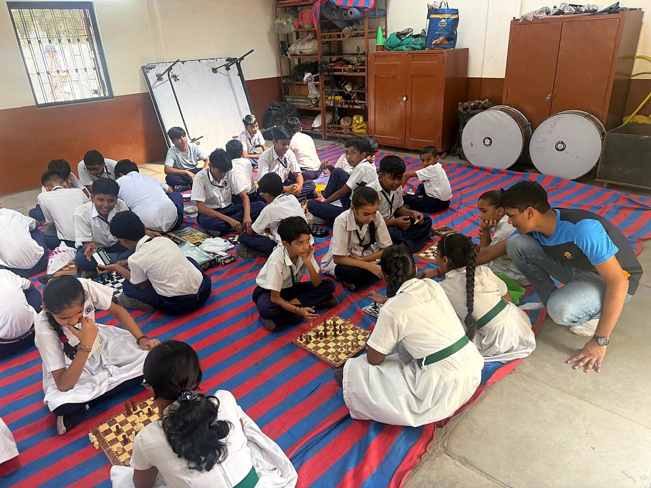 Mukund crouches beside students seated around a chessboard while small groups practise nearby.