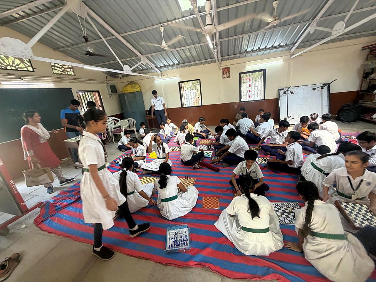 A wide classroom view showing students working in groups around chessboards.
